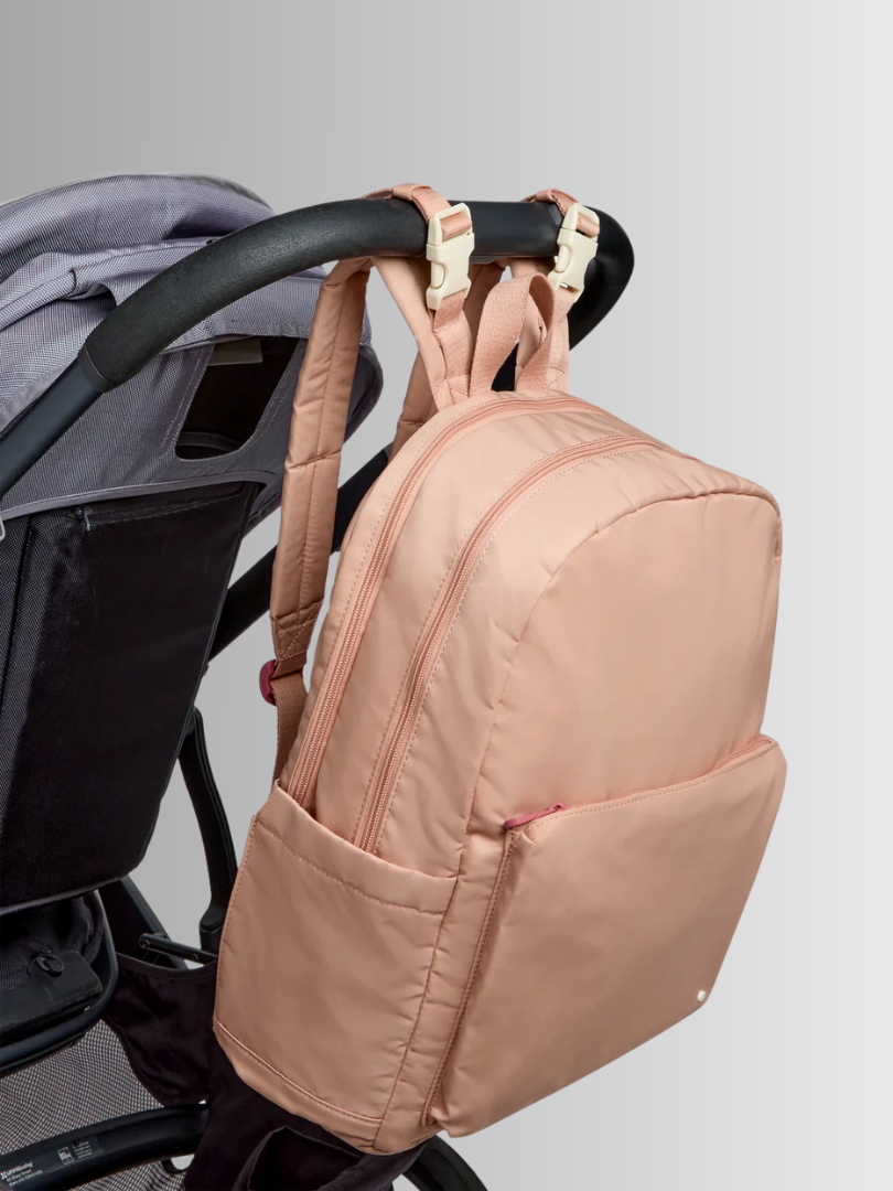 A pink backpack is attached to the handle of a stroller using beige clips, with the stroller partially visible on the left against a light background.