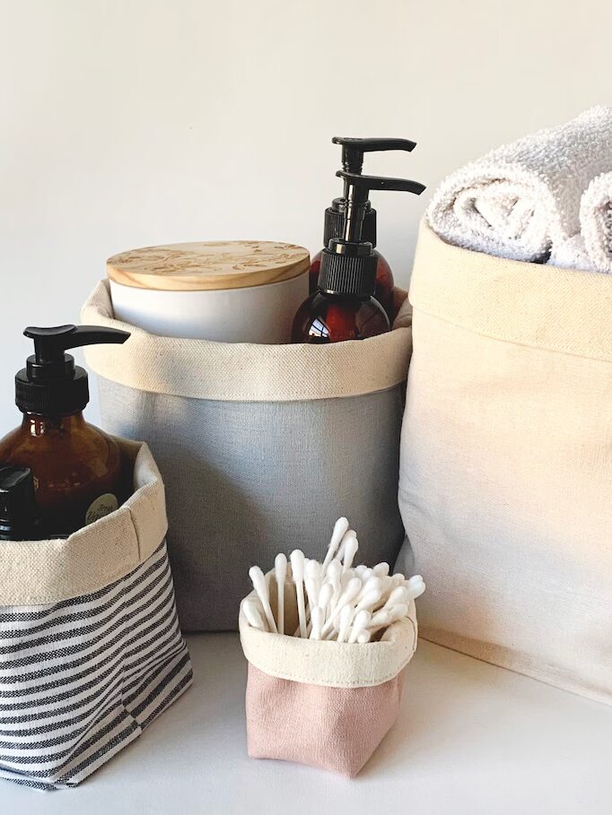 Four fabric storage baskets hold toiletries, liquid soap bottles, cotton swabs, a lidded container, and rolled white towels, all arranged against a plain background.