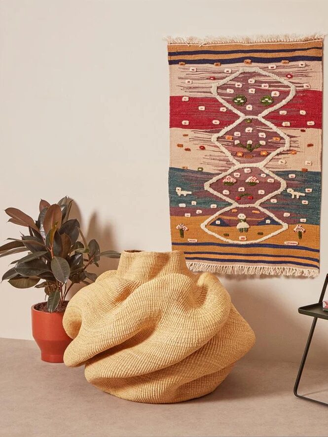 A colorful woven wall hanging with geometric patterns and small animal motifs is displayed above a potted plant, a large knotted cushion, and a black side table with books.