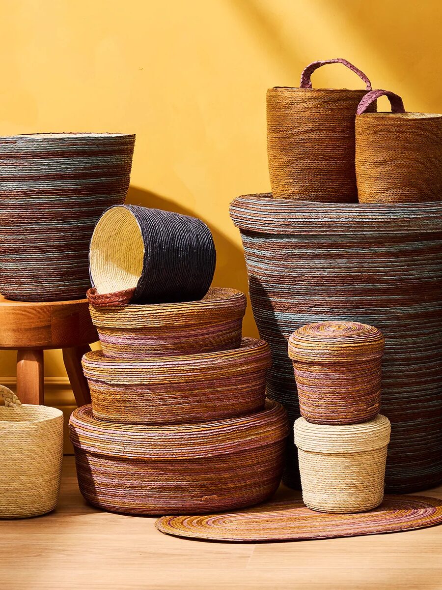 A group of woven baskets in various sizes and shapes, with earthy tones, arranged on a wooden stool and floor against a yellow wall.