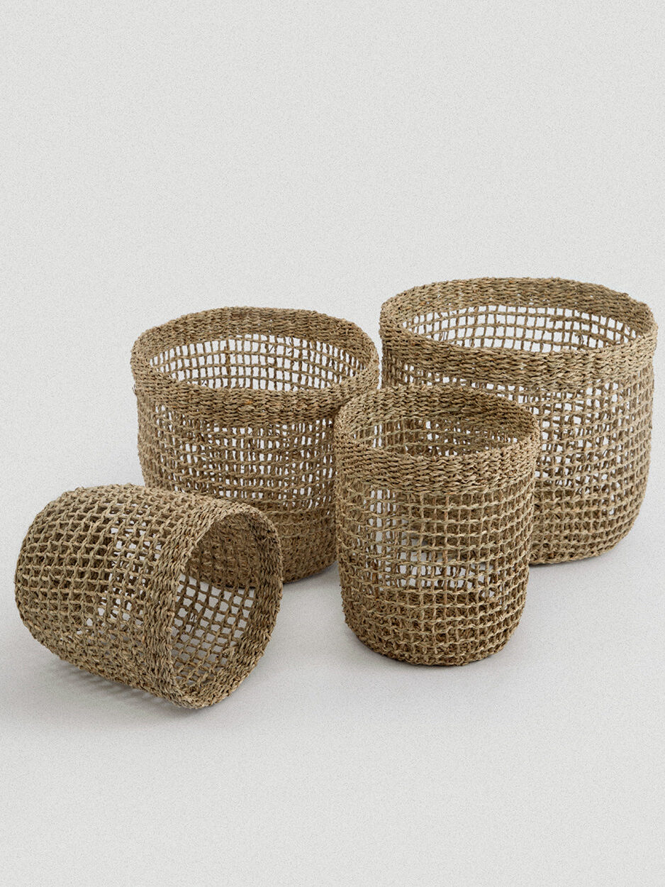 Four woven, round, open-top baskets of varying sizes are arranged on a plain light background. The baskets are made from natural fibers and have a mesh-like design.
