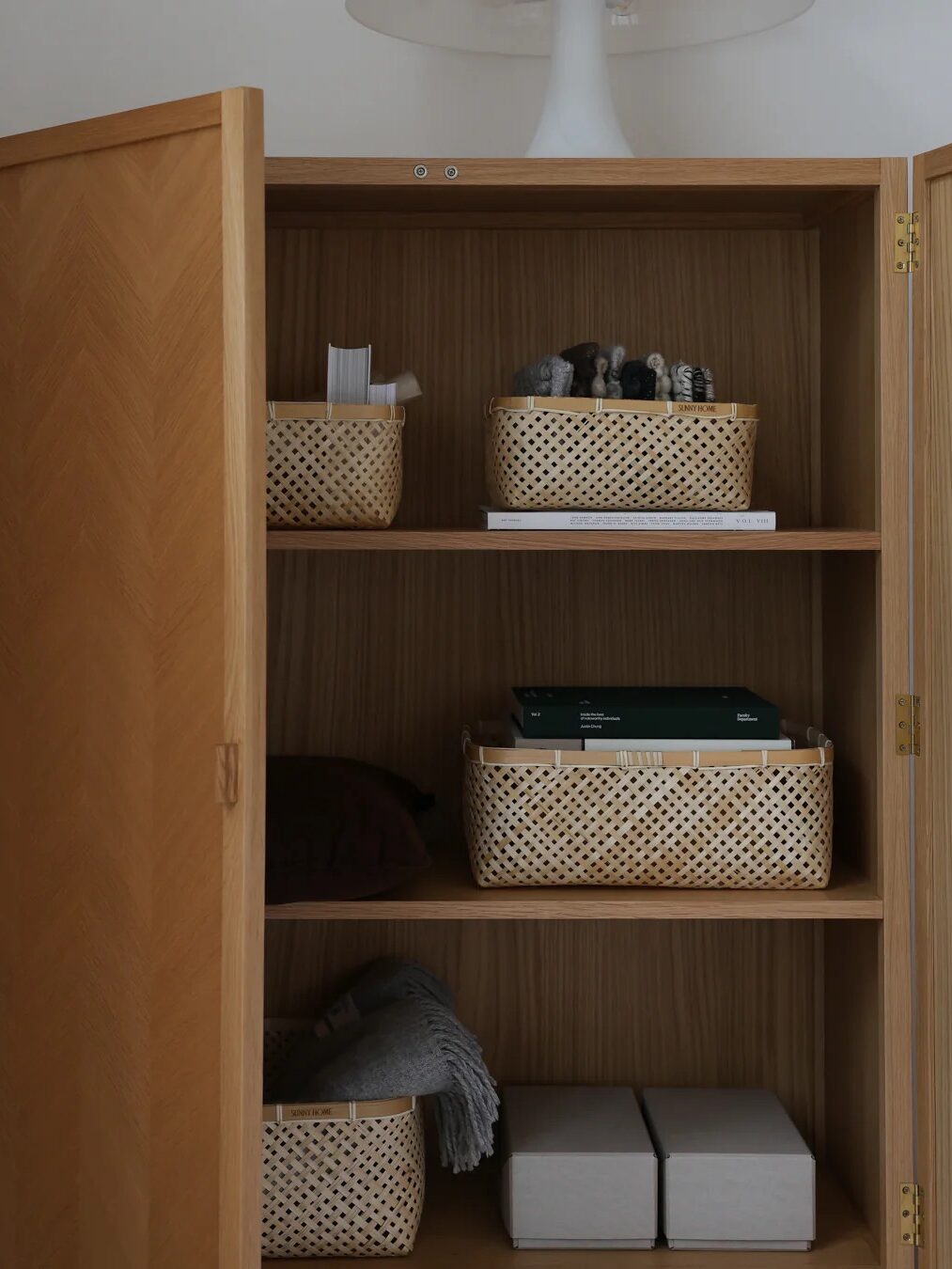 An open wooden cabinet with three shelves holds woven baskets, folded clothes, books, and two gray boxes. A white lamp is partially visible on top.