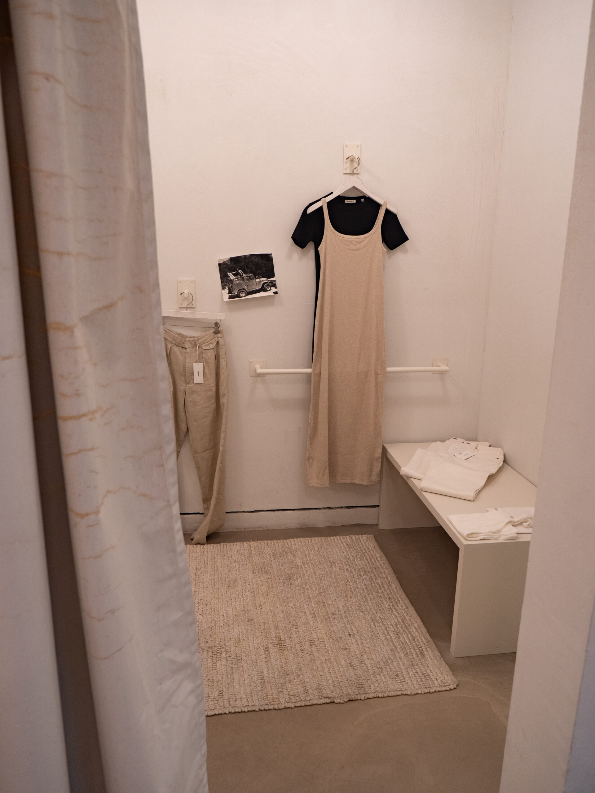 A beige dress layered over a black shirt hangs on a white wall in a minimalist fitting room with beige pants, a rug, a photo, and folded white clothes on a bench.