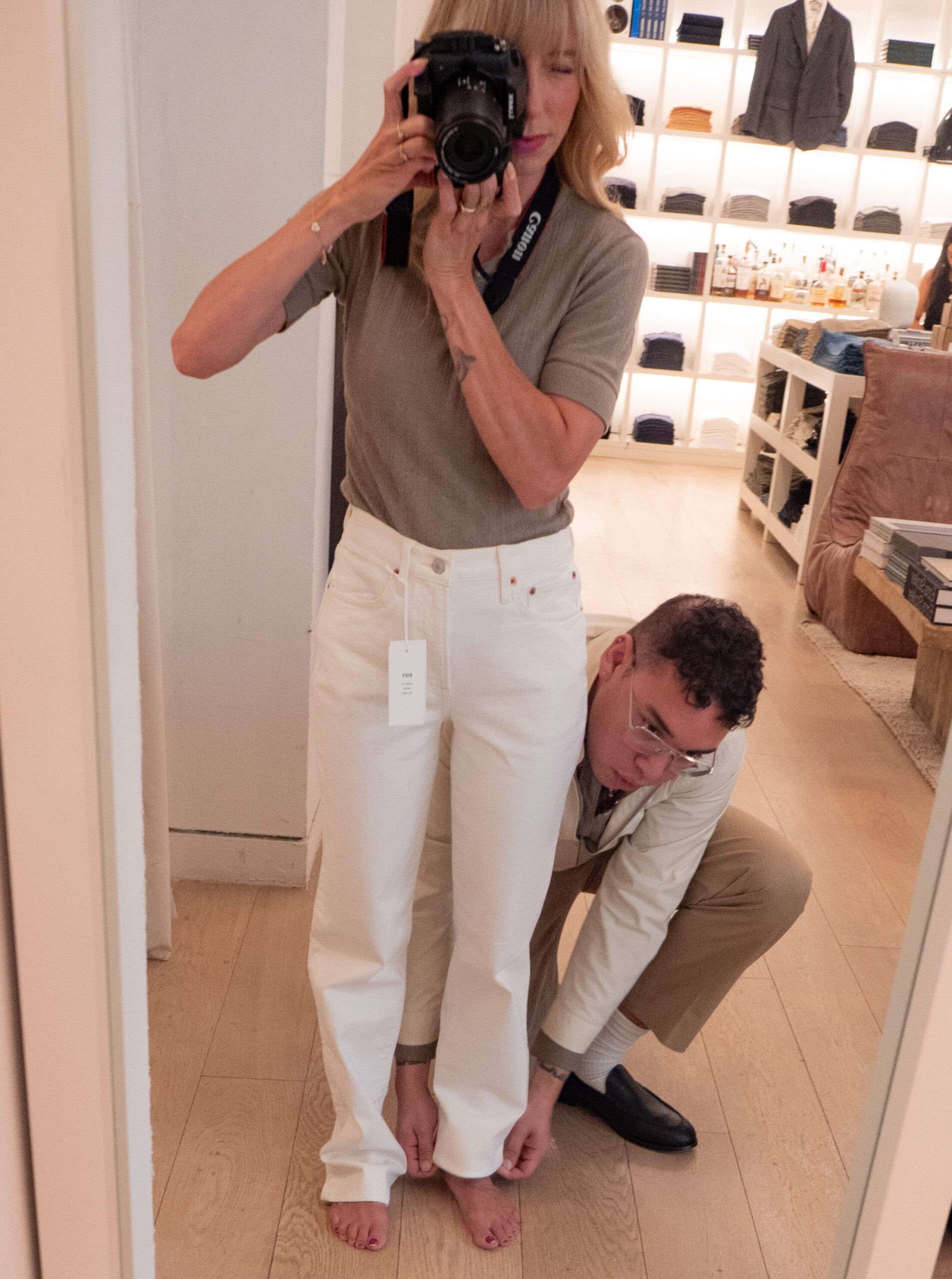 A woman takes a mirror selfie while a man crouches to adjust or examine the hem of her white pants in a store.