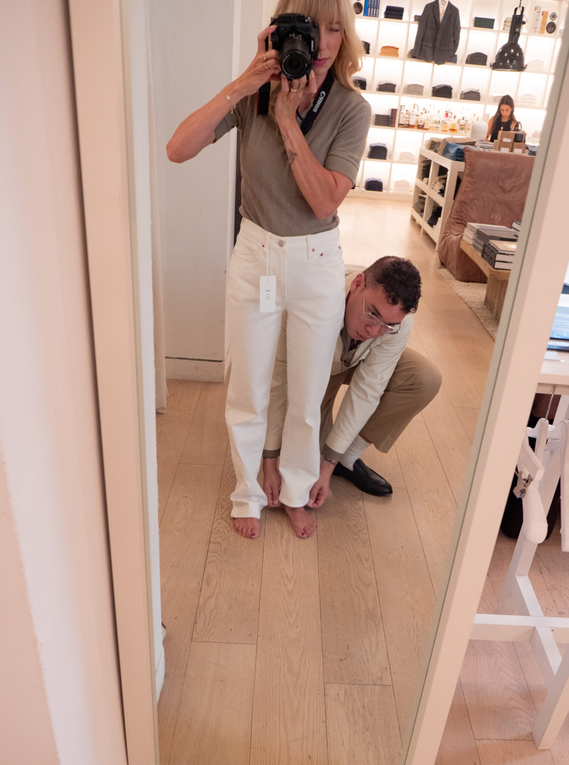A woman takes a mirror selfie while a man crouches by her feet, measuring the length of her white pants in a clothing store.
