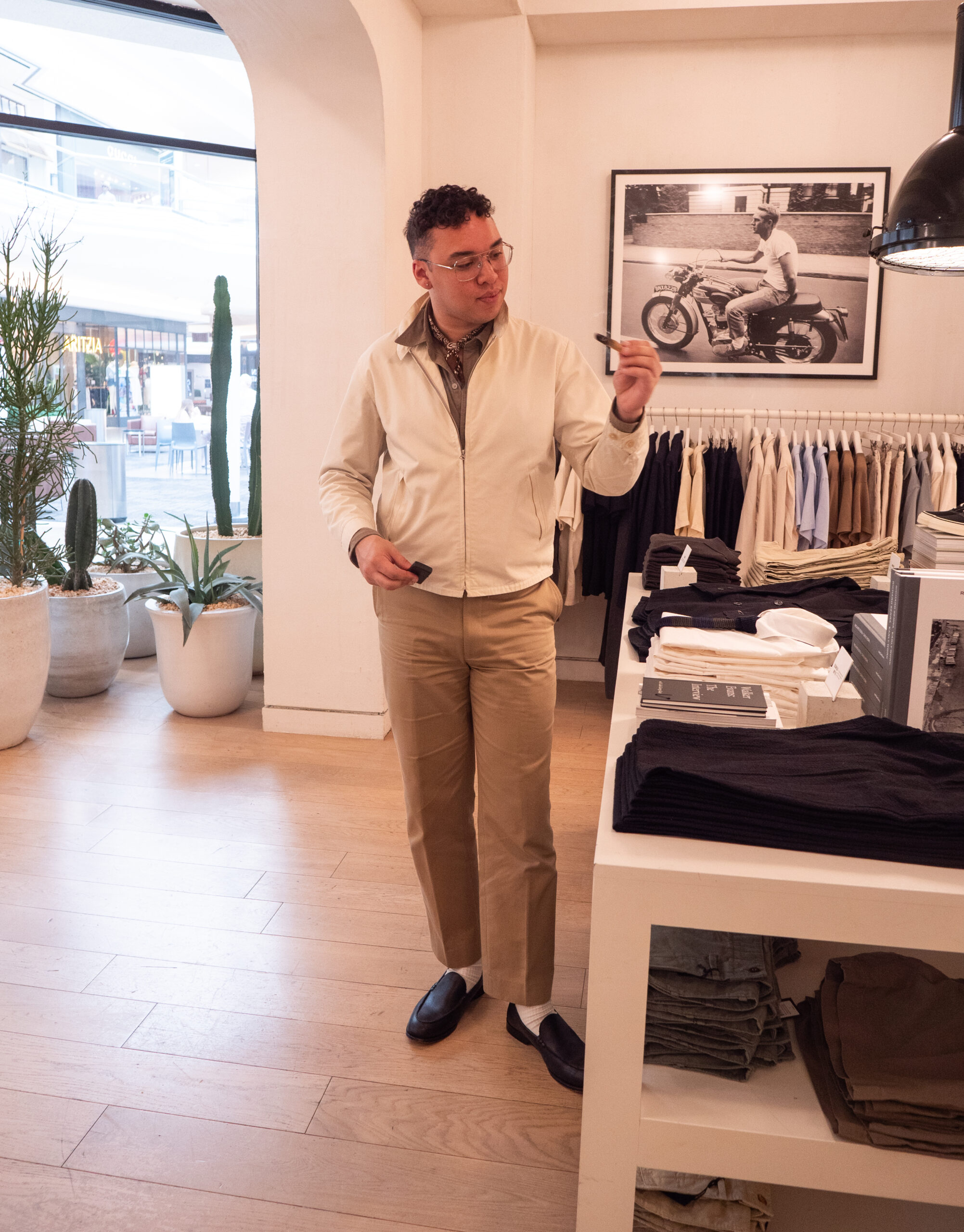 A person stands in a clothing store, holding an item and looking at it. Shelves with folded clothes and racks of shirts are visible in the background.