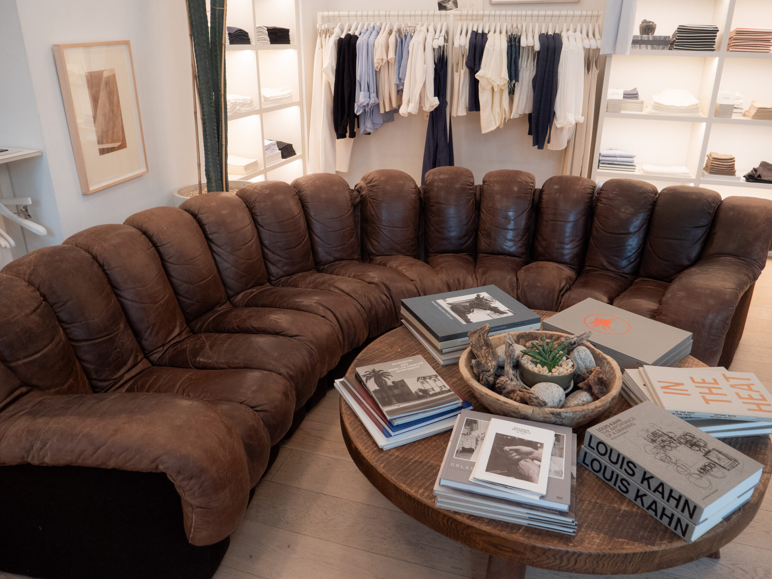 A brown leather sectional sofa surrounds a round wooden coffee table with books and a plant; clothing racks are visible in the background.