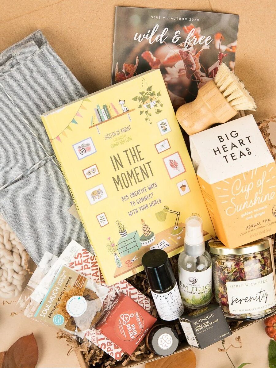 A gift box containing a book, herbal tea, blanket, snacks, a spray bottle, and self-care items is arranged neatly with autumn leaves and a magazine.