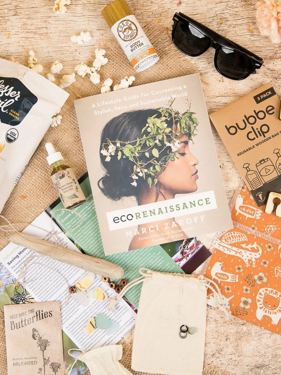 A flat lay of eco-friendly products, including a book titled "ecorenaissance," sunglasses, vegan popcorn, skincare, a reusable bag, plantable paper, and other sustainable items on a wooden surface.