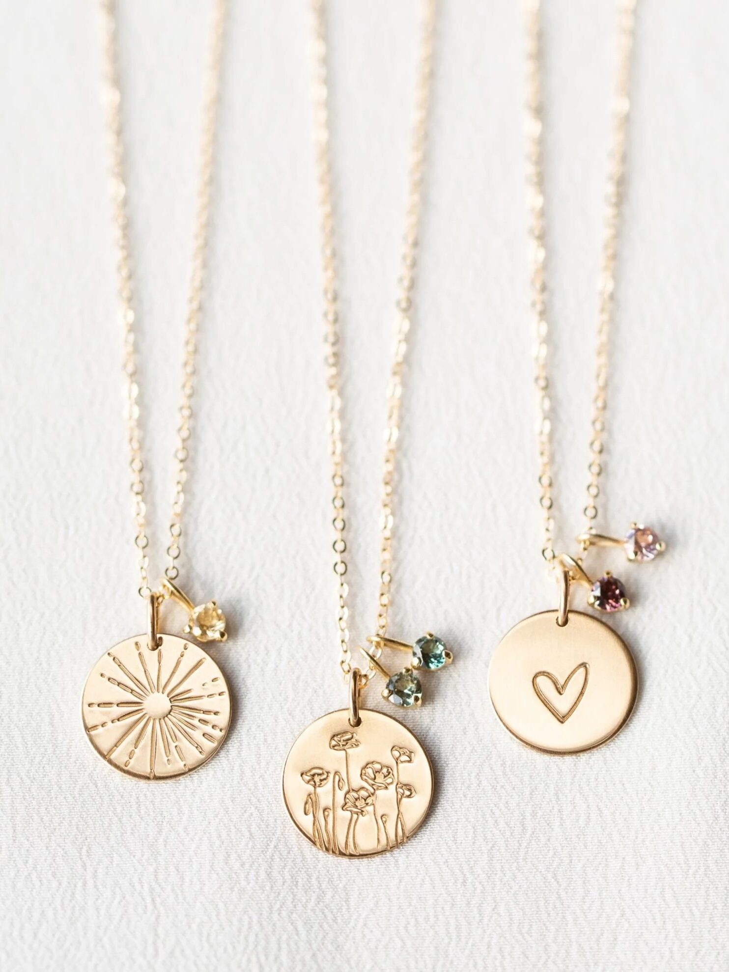 Three gold necklaces with round pendants: one with a sunburst, one with flowers, and one with a heart, each paired with a small gemstone charm, displayed on a white surface.