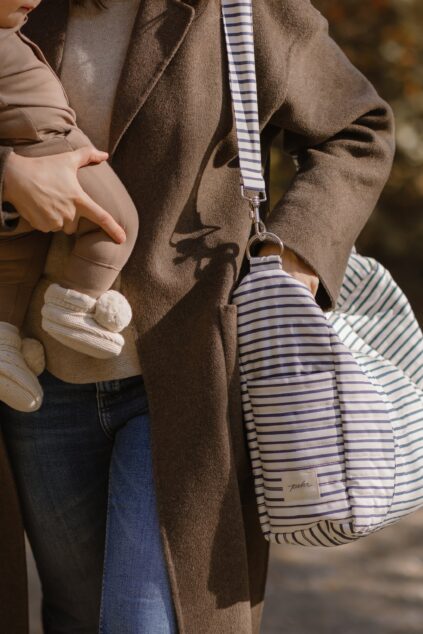 An adult holding a baby wearing beige clothes, with a striped diaper bag and a brown coat visible in the image.