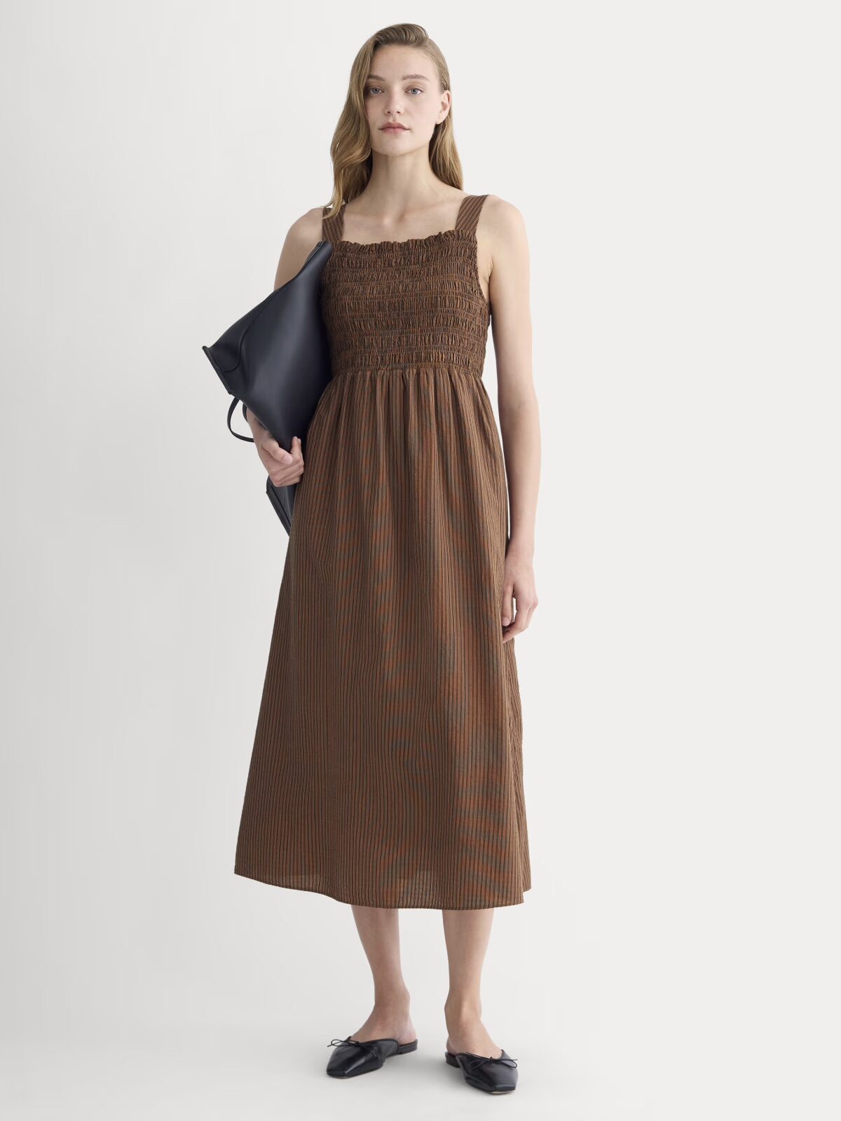 Woman in a brown, sleeveless, smocked midi dress and black flats holding a large black bag, standing against a plain white background.