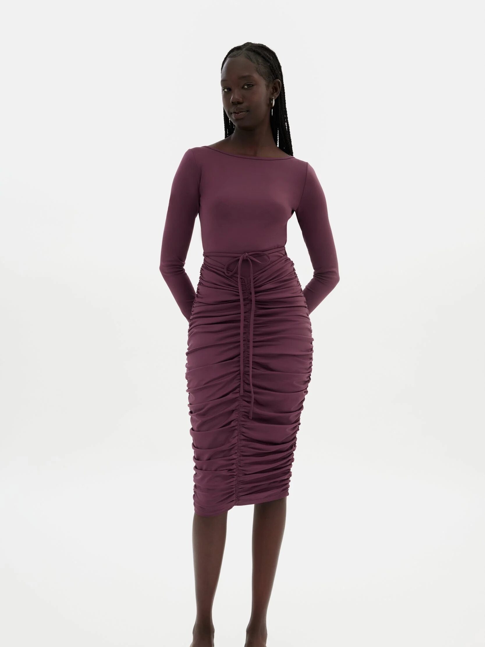 A person wearing a long-sleeve, fitted, ruched maroon dress with a tie detail at the waist, standing against a plain white background.