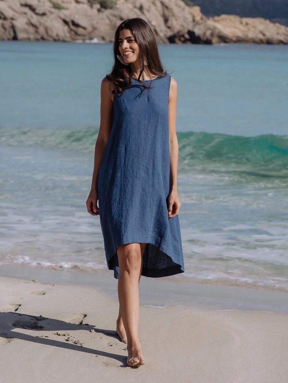 A woman in a sleeveless blue dress walks barefoot along a sandy beach with waves and rocky cliffs in the background.