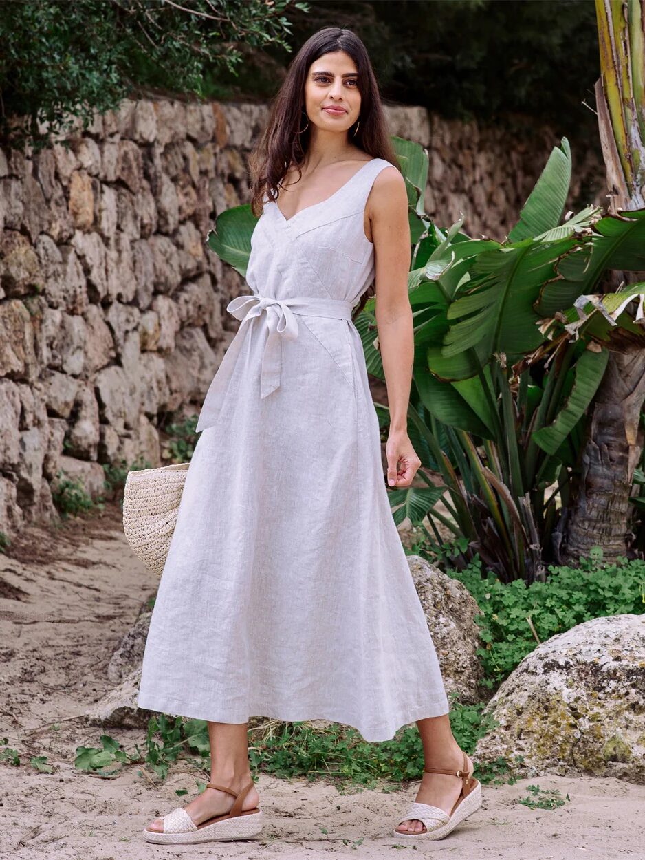 A woman stands outdoors wearing a sleeveless white linen dress with a tied waist, light sandals, and holding a woven bag, near stone walls and tropical plants.