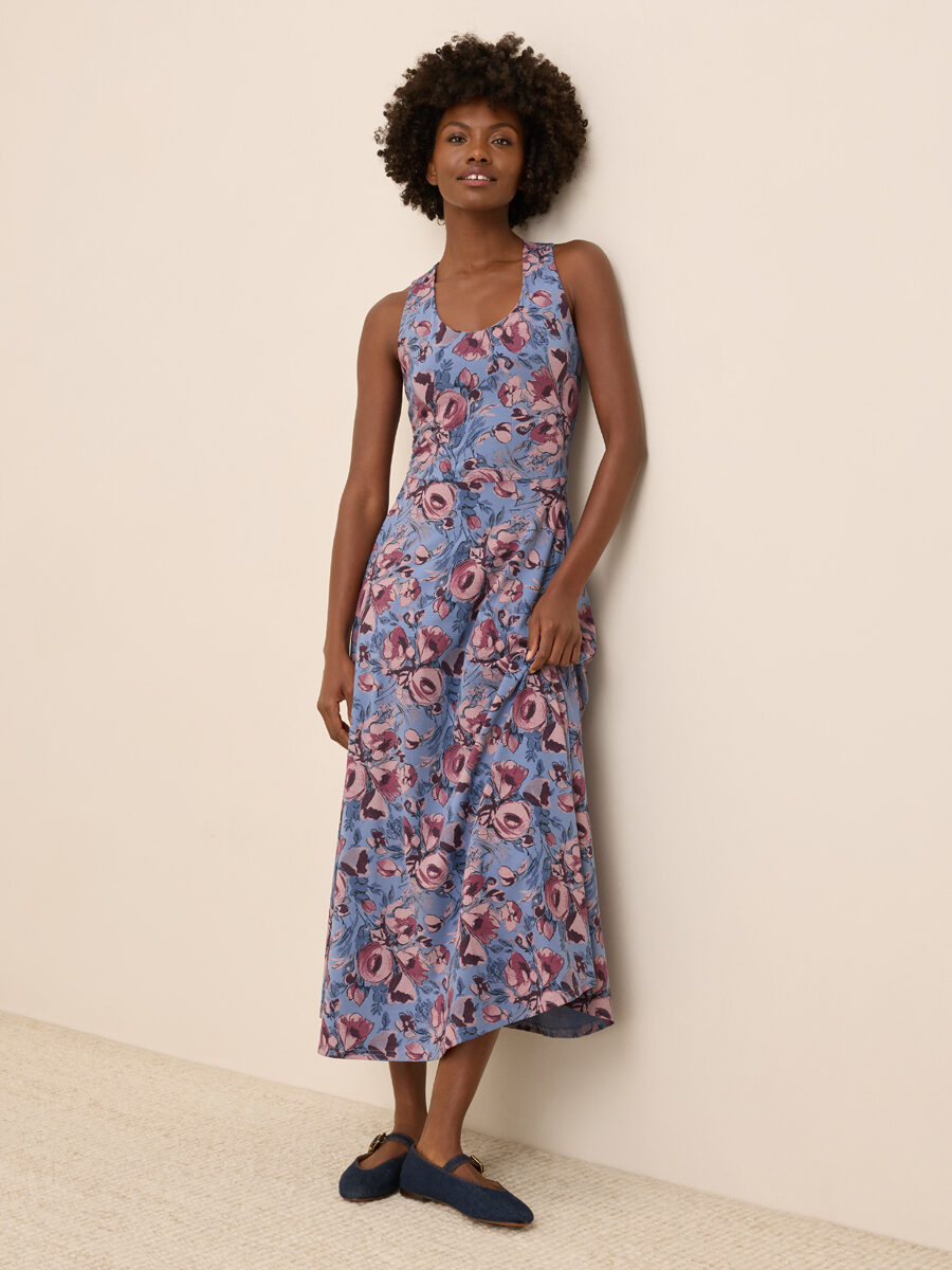 A woman stands against a beige wall wearing a sleeveless, blue floral dress and dark blue slip-on shoes.