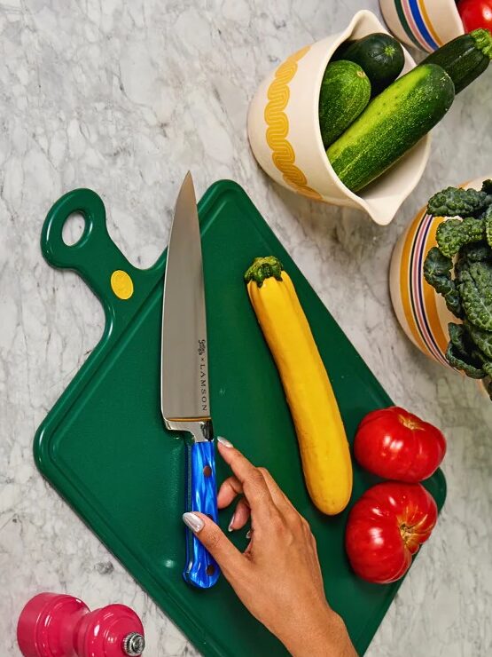 A hand rests near a chef’s knife on a green cutting board with a yellow squash, tomatoes, and various vegetables on a marble countertop.