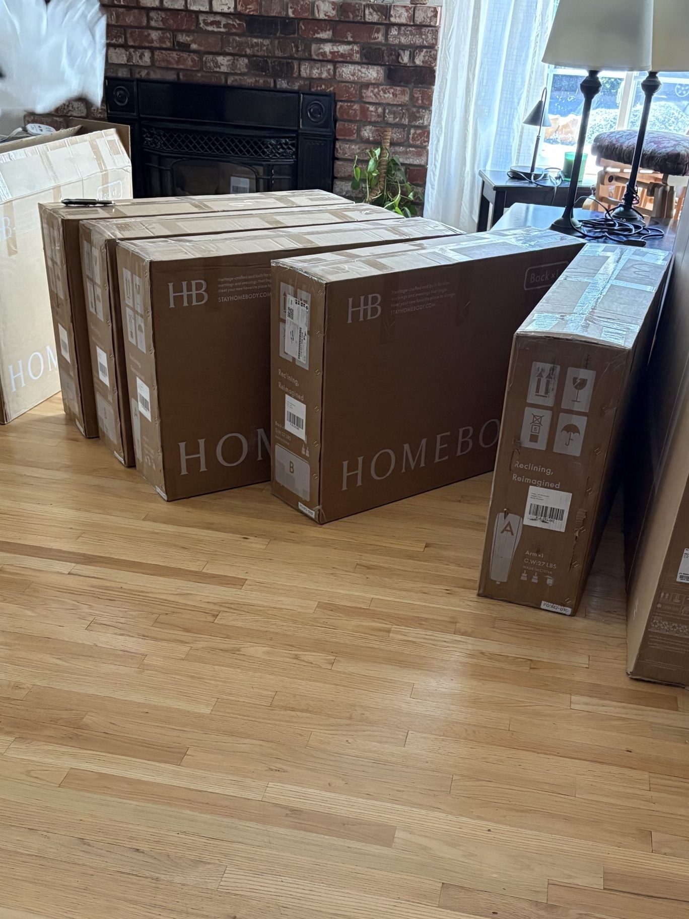 Several large cardboard boxes, labeled "HOMEBOX" and "TCL," are lined up on a wooden floor in a living room near a fireplace. A person wearing gloves stands to the left.