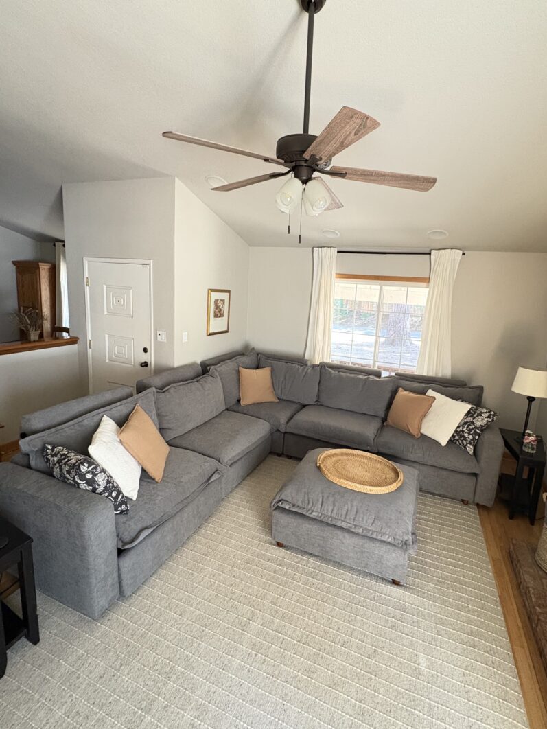 A living room with a large gray sectional sofa, a matching ottoman with a tray, a ceiling fan, a window with curtains, and a brick fireplace on the right.