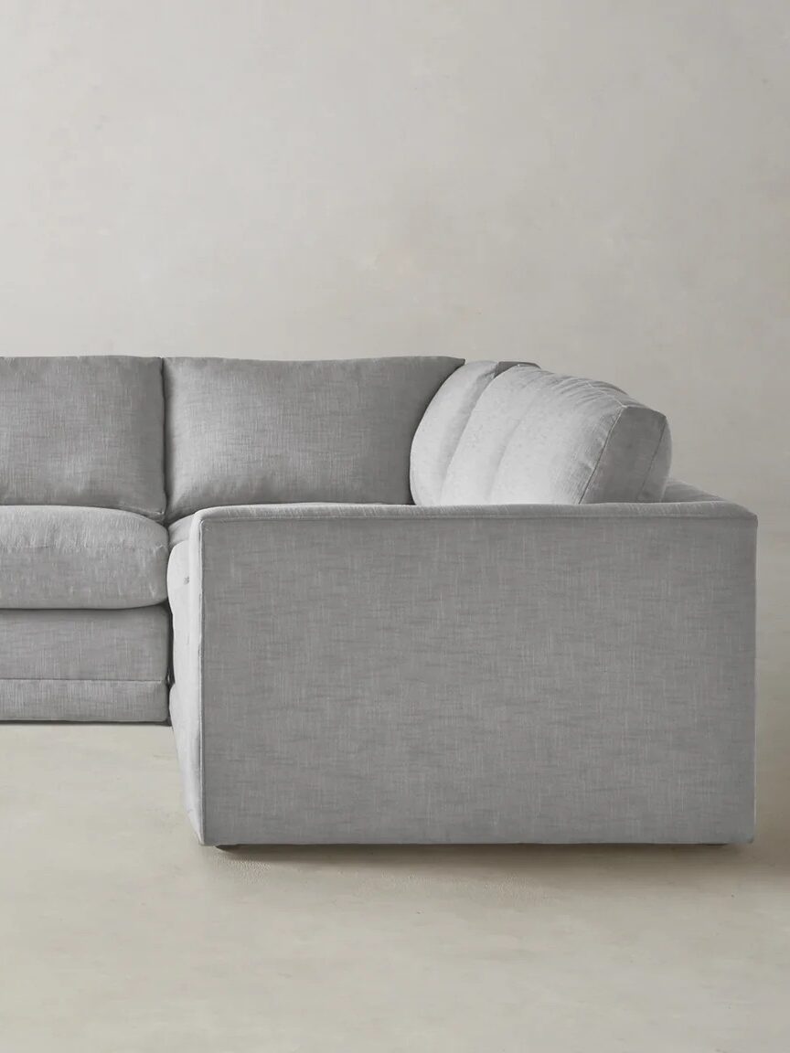 A large, light gray upholstered sectional sofa with clean lines is positioned against a plain light-colored wall and floor.