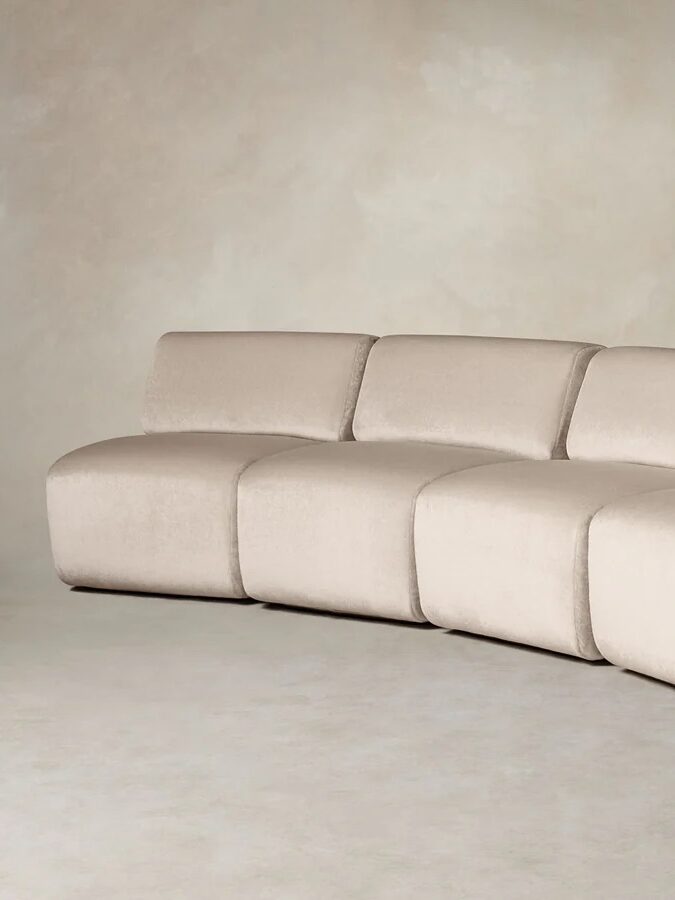 Curved, modular beige sofa with five connected cushioned seats, set against a neutral, textured background.