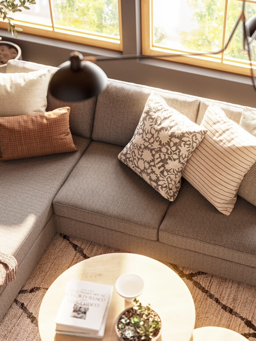 A gray sectional sofa with patterned and striped throw pillows sits by large windows, next to a round wooden coffee table with a book and plant, under modern black lamps.