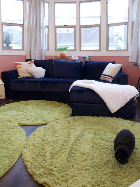 A living room with a navy blue sectional sofa, white blanket, green rugs on dark wood floor, large windows, and a black cat on the rug.