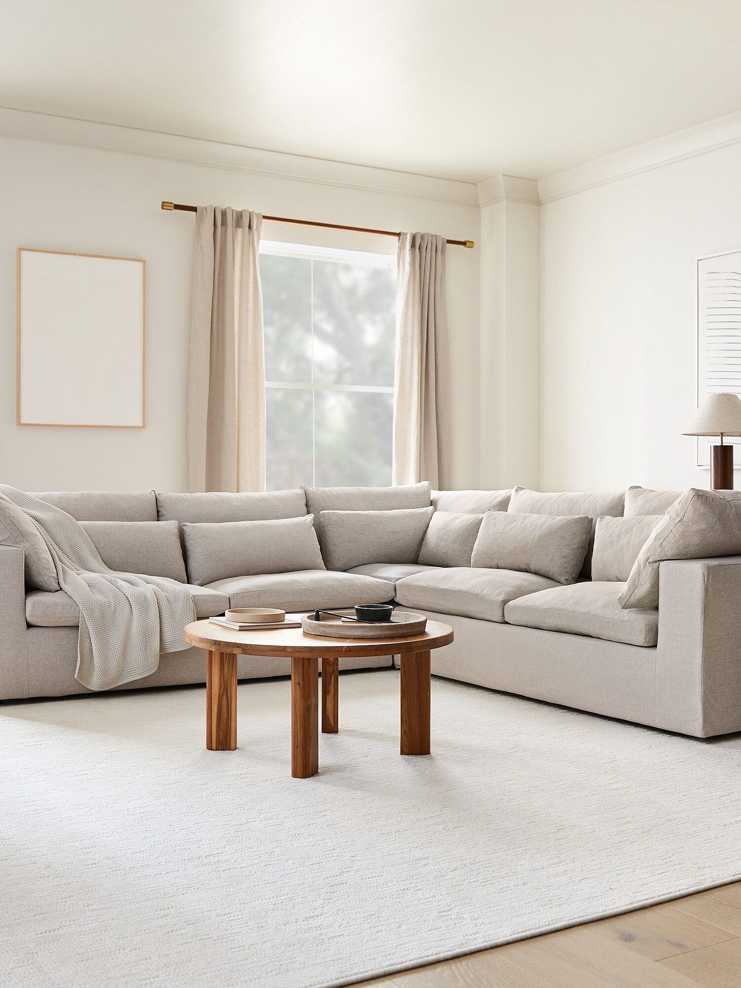 A spacious living room featuring a large beige sectional sofa, two round wooden coffee tables, neutral curtains, and minimal wall art on light-colored walls.