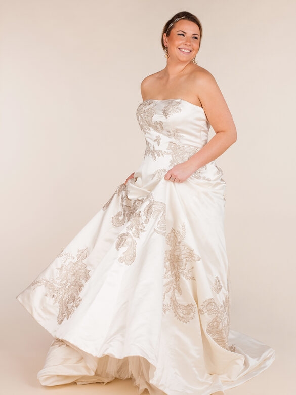 A woman wearing a strapless white wedding dress with embroidered details, standing and smiling against a plain background.