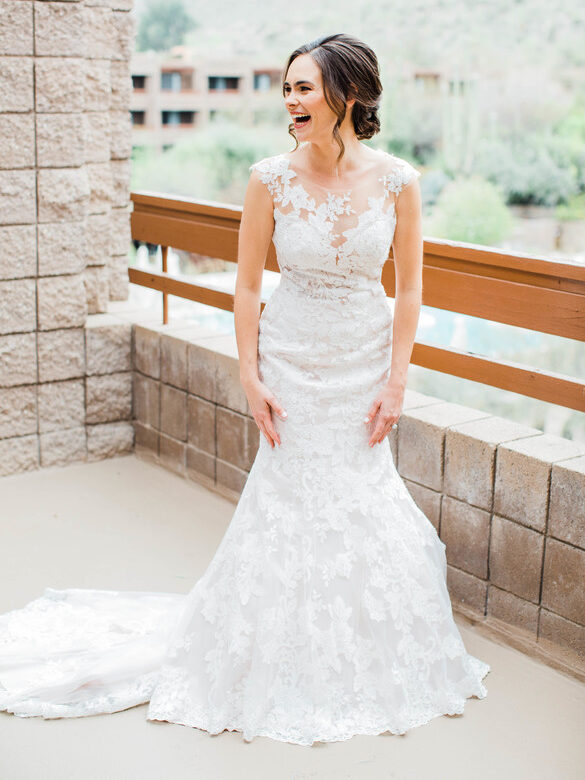 A woman in a white lace wedding dress stands on a balcony, smiling and looking to the side. The background features a stone wall and a blurred outdoor landscape.