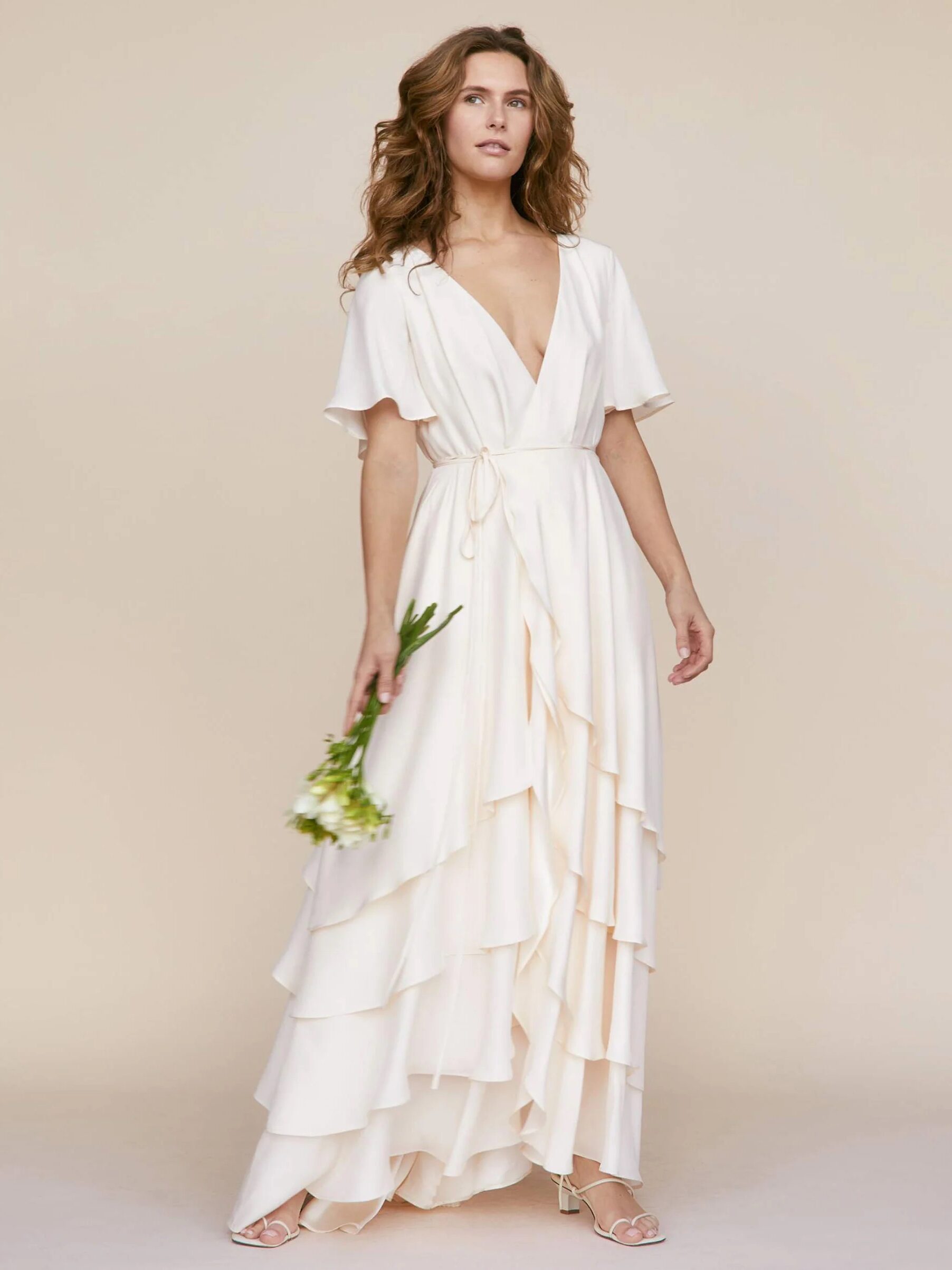 Woman in a flowing, layered white dress holding flowers, standing against a plain beige background.