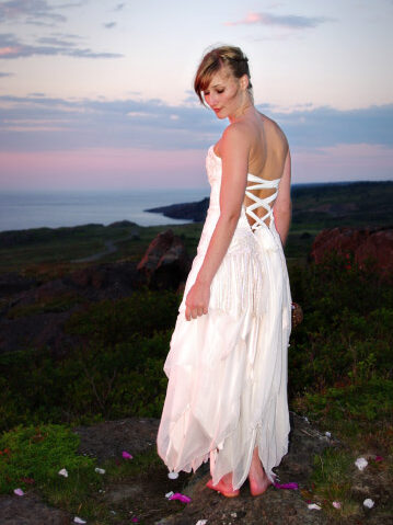 A woman in a white, lace-up back dress stands barefoot outdoors on a rocky area with scattered flower petals, overlooking a scenic landscape at sunset.