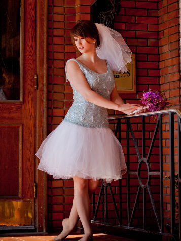 A woman in a short white dress and veil stands on a porch, holding a small bouquet of purple flowers, near a brick wall and wooden door.