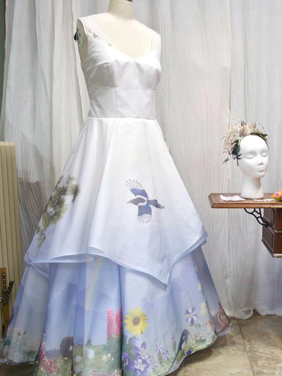 A white sleeveless dress on a mannequin features nature-themed prints, including birds and flowers; a wig with a floral headpiece is displayed on a nearby stand.