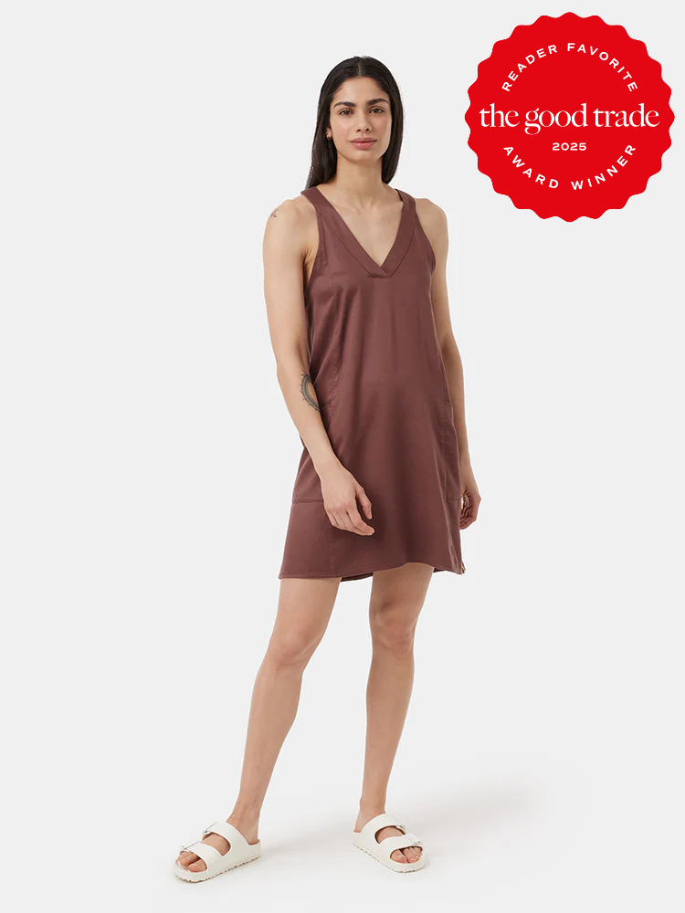 A woman wearing a sleeveless, brown V-neck dress and white sandals stands against a plain background. A red "The Good Trade Award Winner 2025" badge appears in the top right corner.