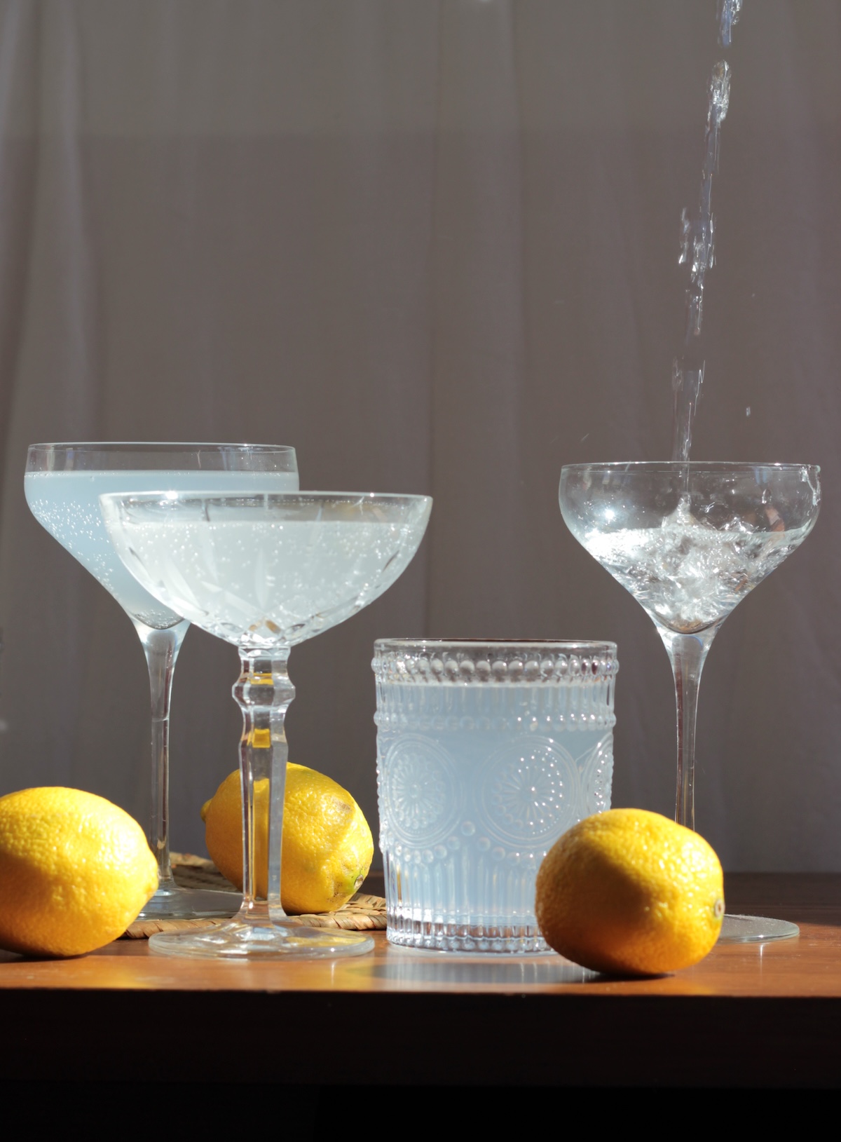 Three glasses filled with a cloudy beverage are surrounded by three whole lemons, with liquid being poured into one of the glasses, set on a dark wooden surface against a neutral background.