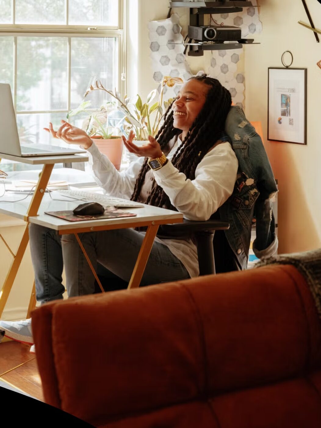 Person sitting at a desk using a laptop, gesturing while on a video call, in a sunlit room with a window, plants, and wall decorations.