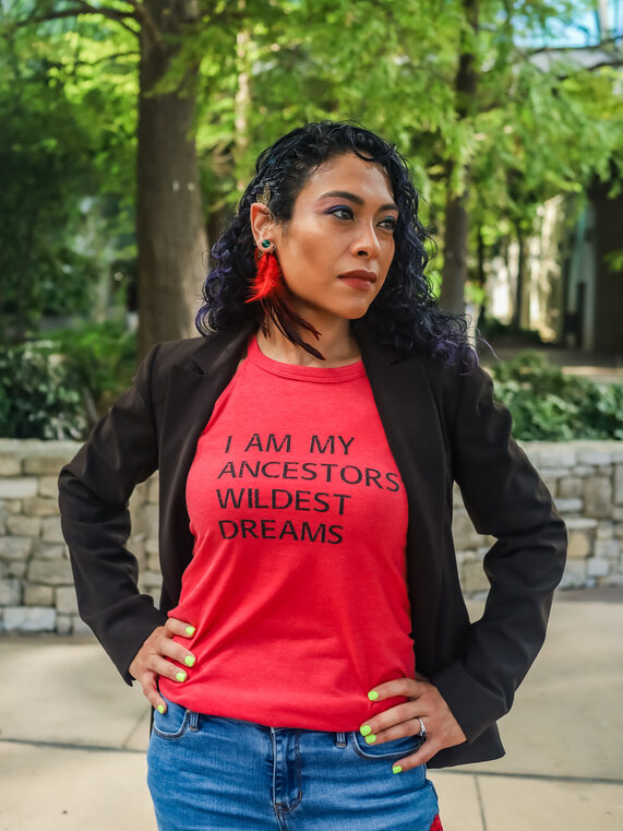 A woman stands outdoors with her hands on her hips, wearing a red shirt that reads "I AM MY ANCESTORS' WILDEST DREAMS," blue jeans, and a black blazer.