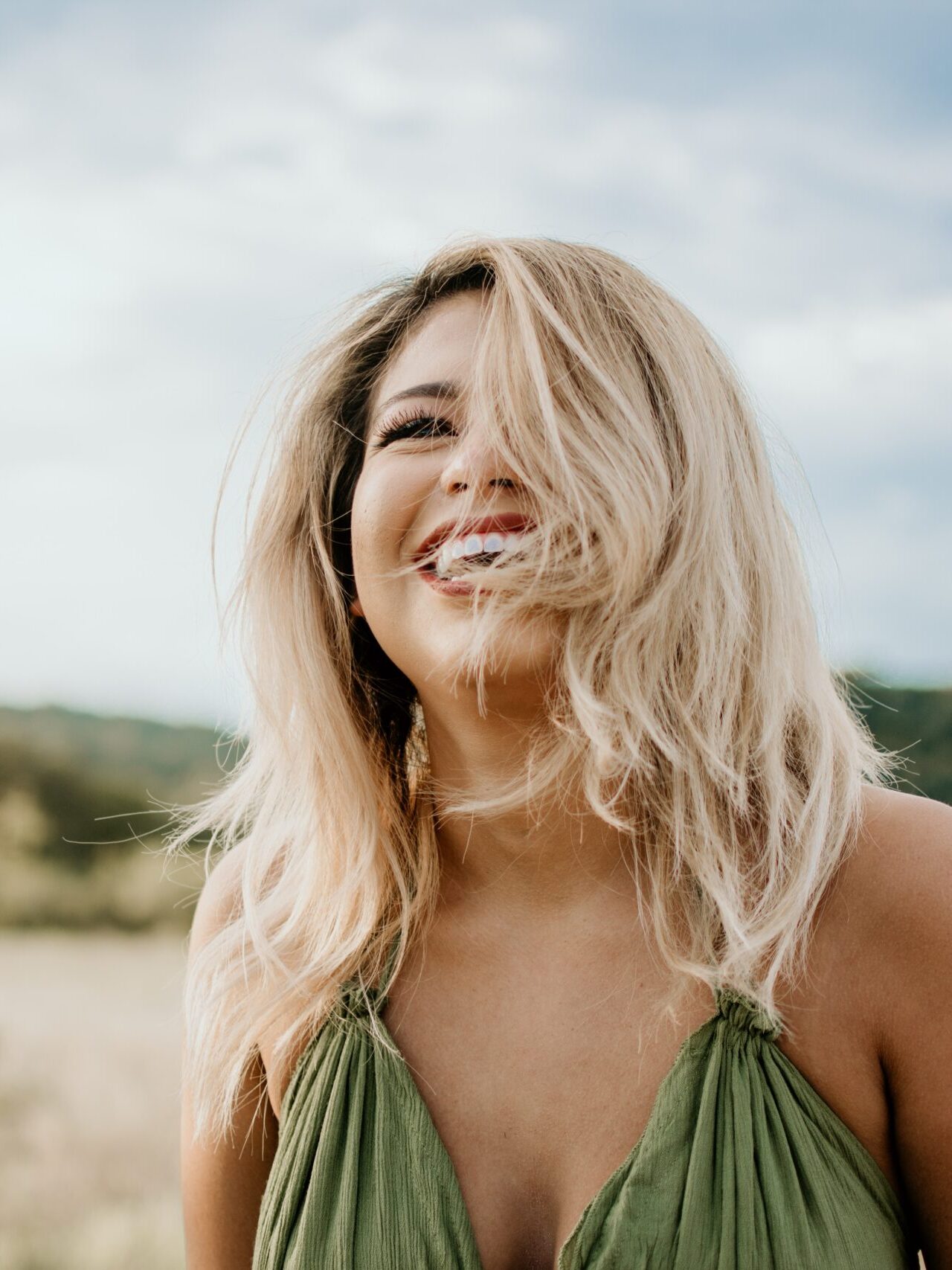 Woman with blonde hair wearing a green dress stands outdoors, smiling with hair partially covering her face, against a blurred natural background.