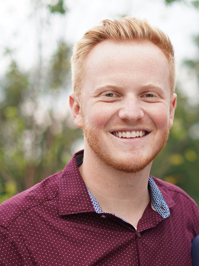 A young man with short red hair and a beard smiles at the camera, wearing a maroon shirt with small white dots. Blurred greenery is in the background.