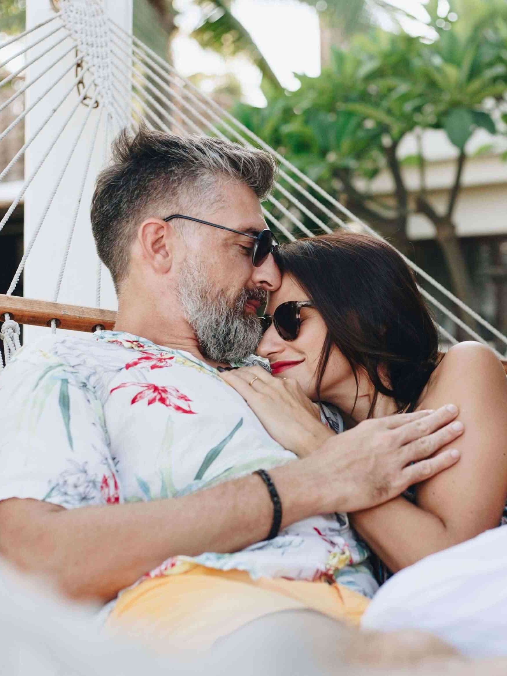 A man and woman wearing sunglasses relax together on a hammock outdoors, surrounded by greenery and buildings in the background.
