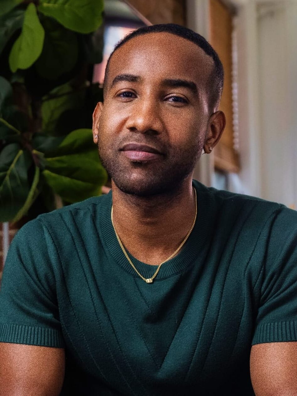 A man with short hair and a trimmed beard sits indoors wearing a green shirt and gold necklace, with a large plant and window in the background.
