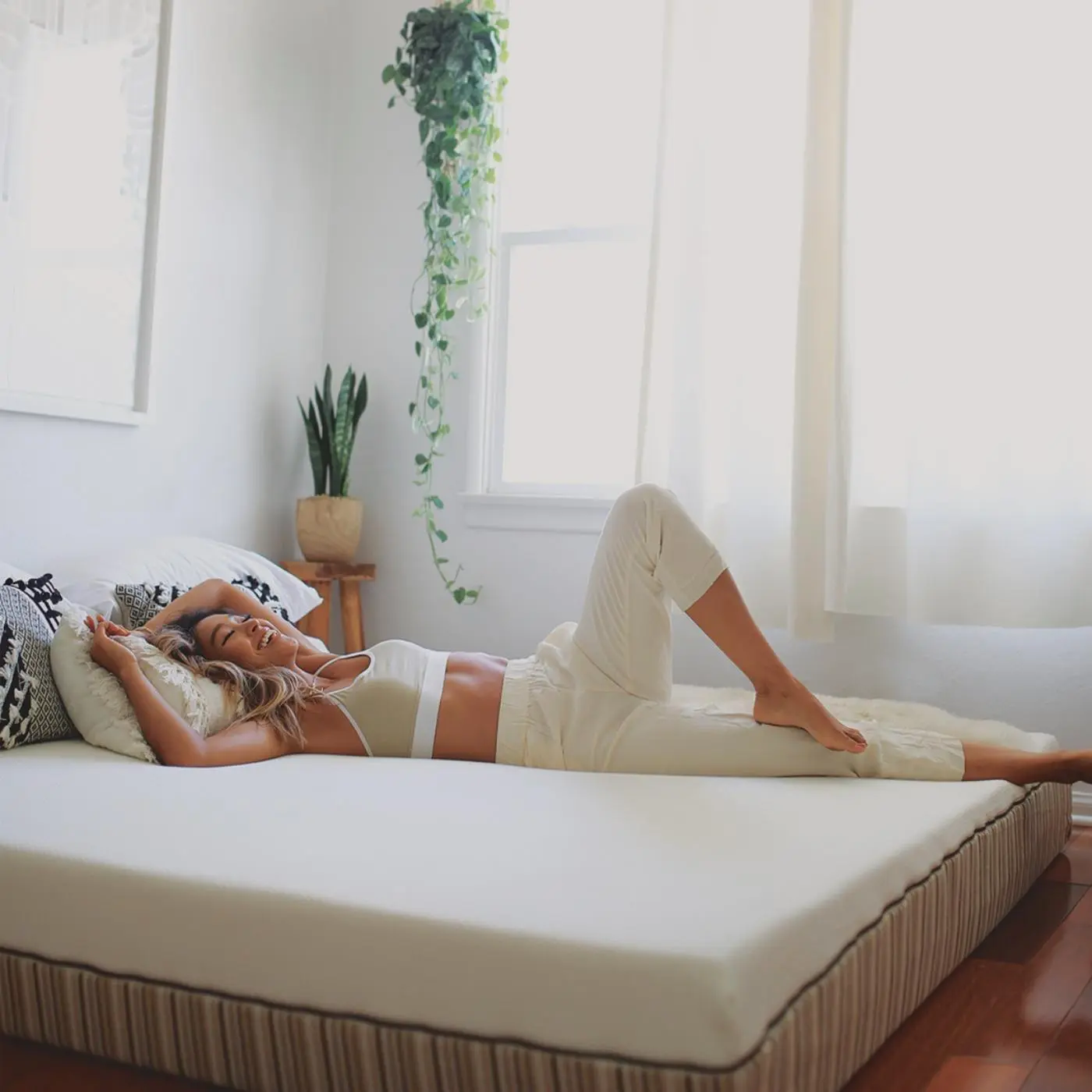 Woman in white loungewear lying on a mattress in a bright room with white walls, a window with sheer curtains, and green plants in the background.