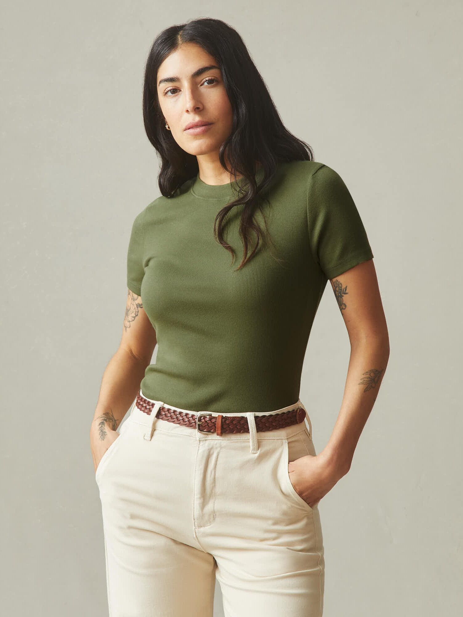 Woman with long dark hair wearing an olive green short-sleeve shirt and light beige pants, standing against a plain light gray background.