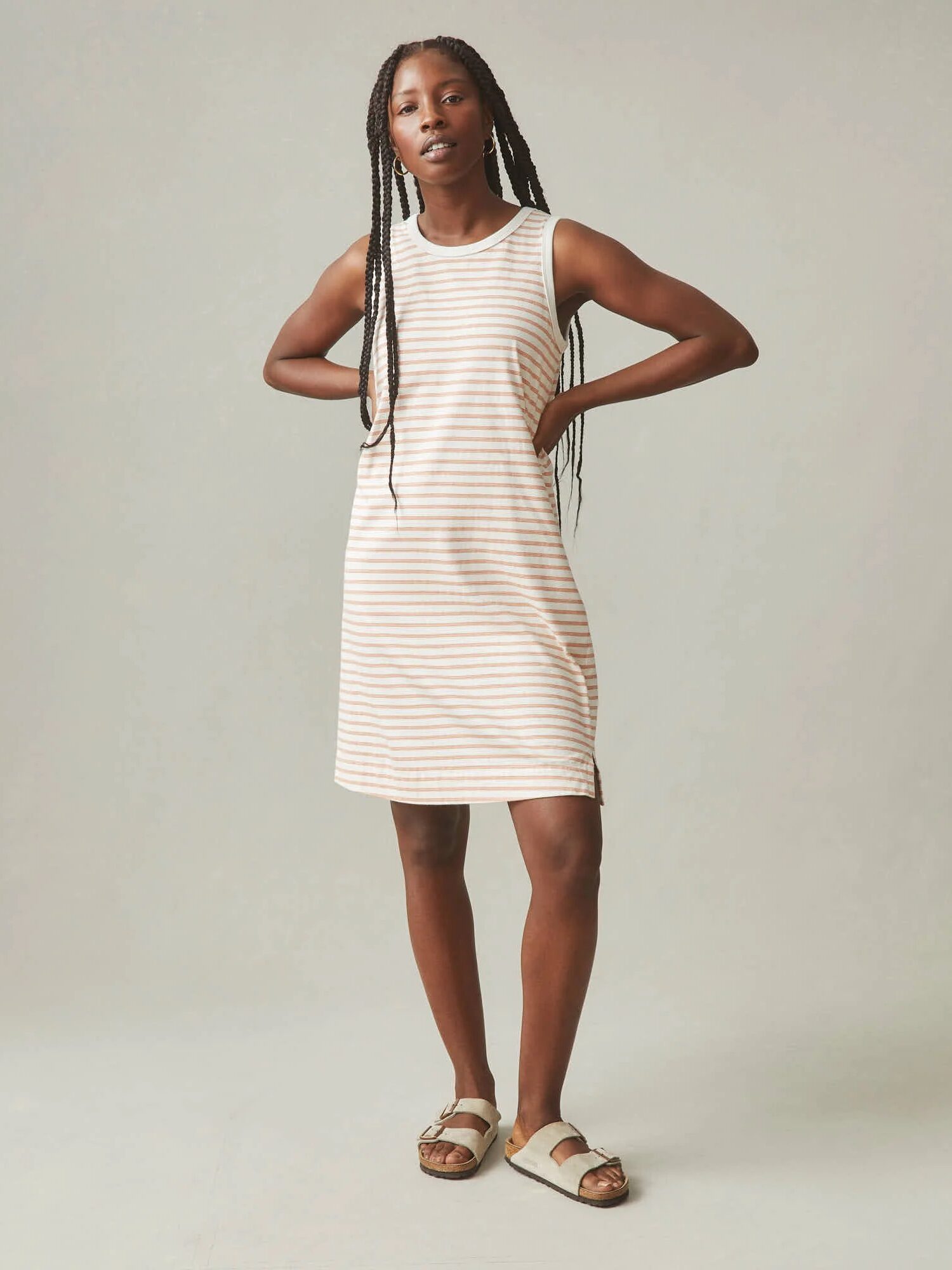 A woman stands with hands on hips, wearing a sleeveless, knee-length striped dress and sandals against a plain background.