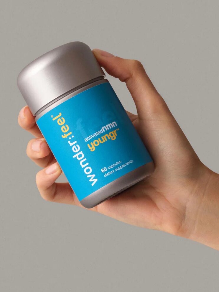 A hand holding a silver bottle labeled "Wonderfeel Younger," containing 60 capsules of dietary supplements, against a plain grey background.
