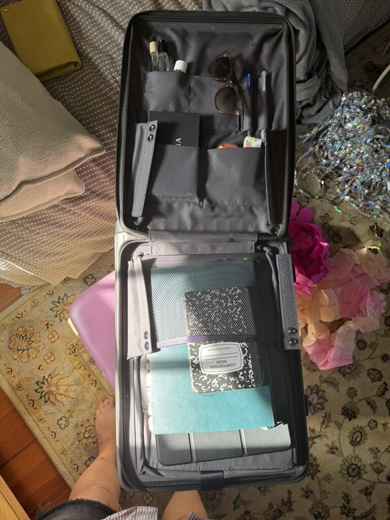 An open suitcase containing notebooks, folders, pens, sunglasses, and other small items sits on a patterned rug with various objects scattered around.