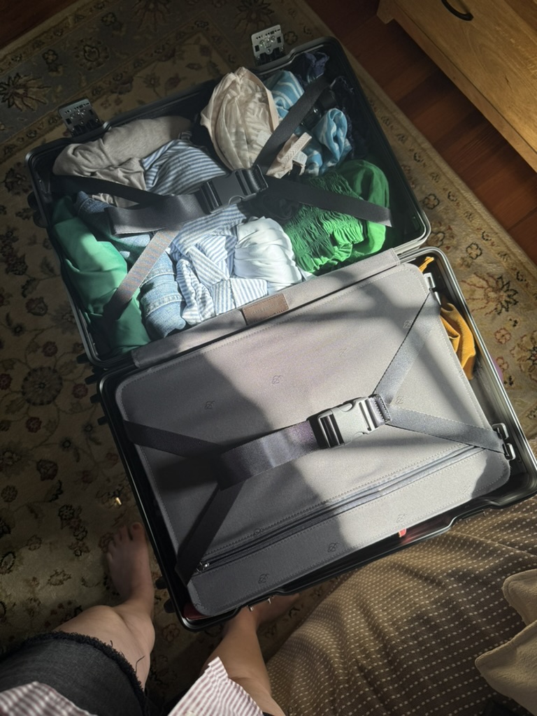 An open suitcase is seen packed with folded clothes and items secured by straps, resting on a patterned rug next to a wooden piece of furniture.