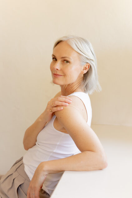 A woman with short gray hair in a white sleeveless top sits sideways, resting her arm on a table and looking at the camera with a slight smile.