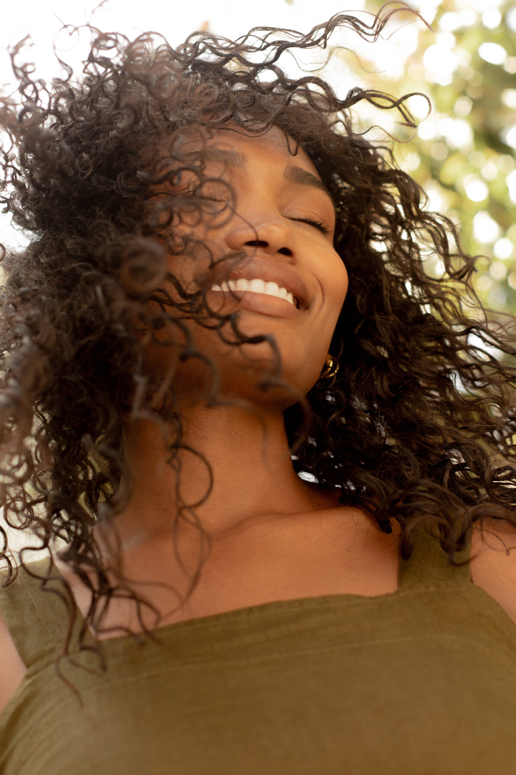 Woman with curly hair smiling with eyes closed, wearing a sleeveless green top, outdoors in natural sunlight.